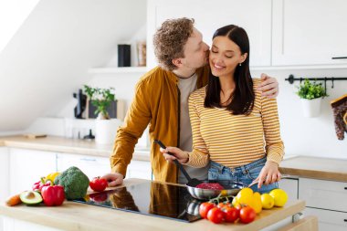 Sevgi dolu bir adam, sıcak mutfakta gülümseyen karısına sarılıp öperken bir kadın, evinde ailesine yemek pişiriyor.