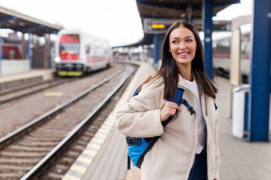Sırt çantalı gülümseyen kadın tren istasyonunda duruyor, trenini bekliyor, bayan hafta sonu gezisine hazır.