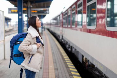 Trenle seyahat et. Sırt çantalı mutlu kadın trene bakıyor, vagonunu kontrol ediyor tren istasyonunda dikiliyor, yandan bakıyor.