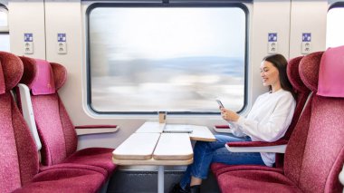 Trende rahat koltukta oturan ve cep telefonuna göz atan, dinlenen, tatilin, manzaranın, manzaranın tadını çıkaran mutlu bir bayan.