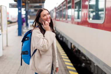 Neşeli bayan gezgin tren istasyonunda cep telefonuyla konuşuyor, tren vagonuna binmek için bekliyor, hafta sonu gezisine hazır.