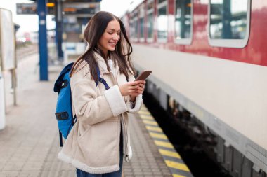 Tren istasyonunda akıllı telefon kullanan genç bir kadın, trenin önünde duruyor, telefonda internet biletini kontrol ediyor.