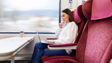 Düşünceli kadın yolcu, tren penceresinin yanında oturuyor ve bilgisayarını uzaktan kumanda olarak kullanıyor. Tren yolculuğu sırasında, yan görüş sırasında çalışıyor.