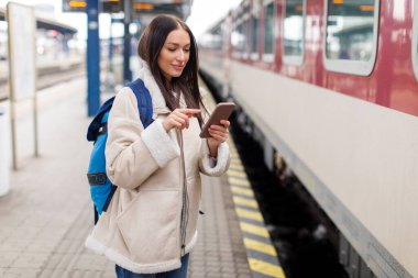 Avrupalı bayan gezgin akıllı telefondan tren tarifesini kontrol ediyor, trenin önündeki platformda duruyor.
