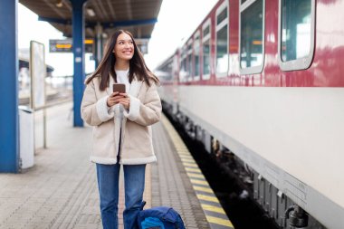 Cep telefonunu kullanan mutlu bir kadın vagon numarasına bakıyor, gülümsüyor, trende bekliyor ve kontrol ediyor, platformda duruyor.