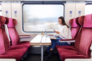 Trenle seyahat ederken ve pencerenin yanında otururken bilgisayarını ve cep telefonunu kullanan pozitif kadın serbest görüşü