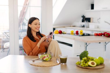 Pozitif genç bayan taze salata yiyor ve cep telefonu kullanıyor. Sağlıklı bir akşam yemeği yerken mutfakta oturuyor.