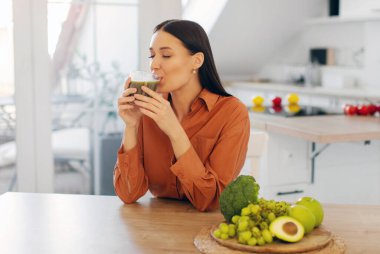 Bardaktan taze detoks suyu içen, masada oturan, evde sağlıklı meyve sularının tadını çıkaran mutlu genç bayan.