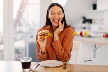 Neşeli bayan lezzetli hamburgerleri yemekten ve gülmekten zevk alır, ellerinde harika burgerler, mutfak içlerindeki masada otururlar, fotokopi çekerler.