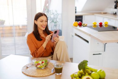 Mutfakta yemek yerken cep telefonu kullanan, bir kase taze salatayla masada oturan ve cep telefonuyla mesajlaşan mutlu bir kadın.