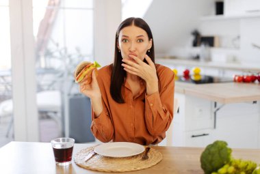 Lezzetli hamburger yiyen, parmağını yalayan ve kameraya bakan, elinde fast food tutan, mutfaktaki masada oturan mutlu kadının portresi.