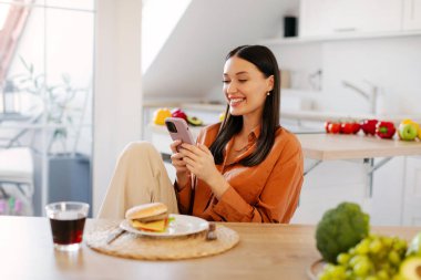 Mutfak masasında oturan mutlu bayan, aldatma yemeği yerken cep telefonu kullanıyor, tabakta burger, bardakta kola. Gadget bağımlılığı ve abur cubur konsepti