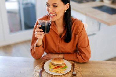 Ellerinde kolayla bardağı tutan ve gülümseyen, mutfaktaki masada oturan, abur cubur yiyen ve ev yapımı hamburger yiyen mutlu kadın.
