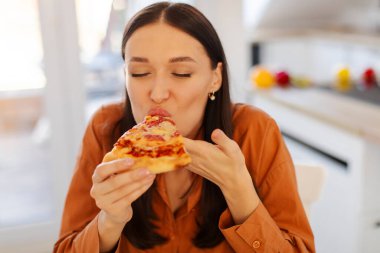 Pizza tutan ve ısıran bir kadının portresi, mutfak içi, yakın çekim. Abur cubur aşığı, sağlıksız beslenme aldatma yemeği