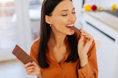 Çikolata yiyen heyecanlı bir bayan, kapalı gözlerle leziz tatlıların tadını çıkarıyor, mutfak içi, yakın plan, yukarısı. Sağlıksız beslenme, aldatma yemek konsepti