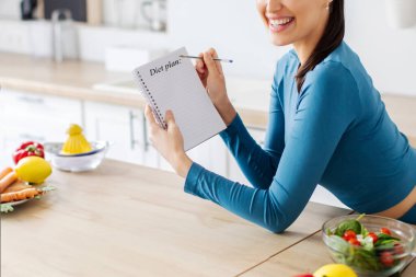 Gülümseyen sporcu kadın menü yazıyor ve deftere sağlıklı kilo verme tarifleri yazıyor, modern mutfakta duruyor. Zayıflama beslenme kavramı