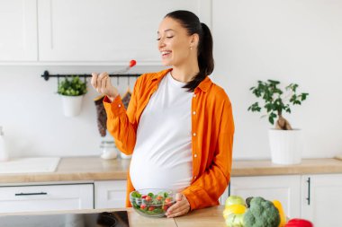 Mutlu hamile bir kadın, şık mutfağında taze sebze yerken neşeli bir şekilde gülümser. Hamilelik boyunca sağlıklı beslendiğini vurgular, fotokopi çeker.