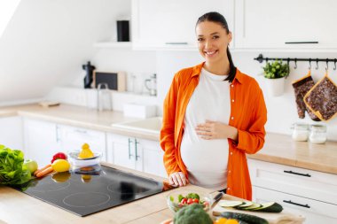Sağlıklı yemek hazırlarken hafif mutfakta duran neşeli hamile kadın koca göbeğine dokunuyor. Sağlık, diyet ve yararlı beslenme
