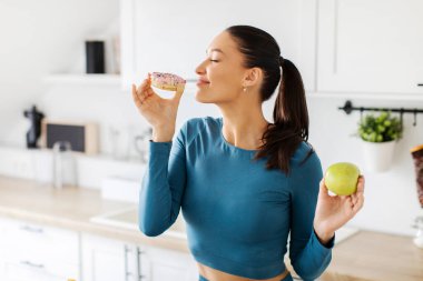 Spor giyim sektöründe formda bir bayan sağlıklı elma yerine şekerli çöreği seçiyor, mutfakta durup lezzetli çörekleri kokluyor. Yemek seçimi ve sağlıklı beslenme alışkanlığı