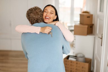 Heyecanlı bir kadın taşınma gününde kocasına sarılıyor ve ev anahtarını tutuyor. Eşler taşınma sırasında yeni evlerinde kucaklaşıyor.