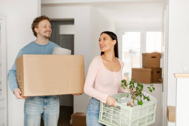 Taşınma günü. Karton kutu ve bitki taşıyan mutlu eşler, eve taşınmak, eşyaları taşımak ve birbirlerine gülümsemek.