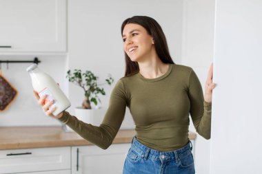 Genç bir kadın buzdolabından bir şişe süt alıyor ve gülümseyerek yanına uzanıyor.