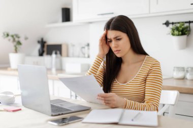 Stresli bayan girişimci evrak işleriyle uğraşıyor ve dizüstü bilgisayarla çalışıyor, mutfakta masada oturuyor, mali belgeleri kontrol ediyor ve kafa kafaya veriyor.