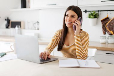 Cep telefonuyla konuşan, dizüstü bilgisayar kullanan, evde çalışan, masada oturan ve gülümseyen pozitif kadın serbest yazar.