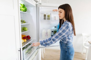 Kadın buzdolabının çekmecesini açıyor. Taze salata ve yemek hazırlamak için ürünleri çıkarıyor. Evde, yan görüşte malzemeler arıyor.