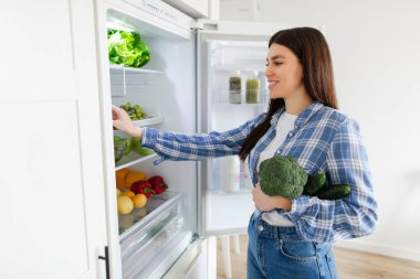 Dolaptan taze yeşil sebzeler alan bir kadın yemek hazırlamak için sağlıklı yiyecekler topluyor.