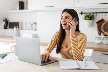 Evden çalışan, cep telefonuyla dizüstü bilgisayarın önünde konuşan, mutfaktaki masada oturan mutlu Avrupalı kadın serbest yazar.