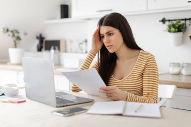 Stresli kadın serbest çalışan, finansal belgelerle çalışan, dizüstü bilgisayarlara bakan, sorun yaşayan, kafa karıştıran, mutfaktan çalışan