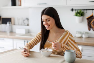 Pozitif kadın kahvaltı yapıyor ve cep telefonu kullanıyor, mutfaktaki sosyal medyada sörf yapıyor, yulaf ezmesi yiyor ve blog okuyor.