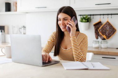 Uzaktan kumandalı kariyer. Cep telefonuyla konuşan ve mutfakta bilgisayar kullanan genç bir bayan. Evde uzaktan çalışan bir serbest çalışan.