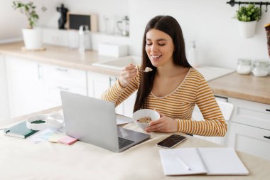Kahvaltısını yapan ve dizüstü bilgisayar kullanan pozitif bir kadın, evde bilgisayardan giyinen kadın, boş yer.