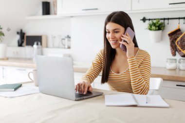 Mesafe. Mutlu bayan serbest çalışan cep telefonuyla konuşuyor ve mutfakta dizüstü bilgisayar kullanıyor, bilgisayarla çalışıyor ve cep telefonuyla sohbet ediyor.