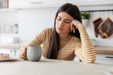Kahve fincanı olan yorgun bir kadın mutfaktaki masada uyuyor, gözleri kapalı oturuyor. Evde fazla çalışma, sabahın erken saatleri ve tek başına kahvaltı.