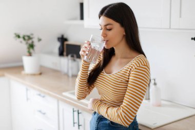 Camdan temiz maden suyu içen genç bir kadın. Mutfağın içinde duruyor. Sağlıklı içecek, vücut suyu konsepti.