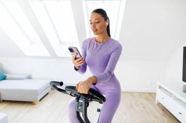 Spor kıyafetli genç bir kadın durağan bisiklette egzersiz yapıyor ve cep telefonunu kullanıyor, müzik arıyor, evinde hafif bir oturma odası var.