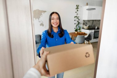 Mutlu bayan müşteri kurye ellerinden paket kutusu alıyor, kapıda duruyor ve gülümsüyor, lojistik hizmeti için teslimatçıya teşekkür ediyor.