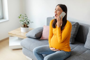 Stresli hamile kadın karnına dokunuyor ve acil durum çağrısı yapıyor, bağırıyor, karın ağrısı çekiyor, kanepede oturuyor, boş yer var.