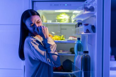 Pijamalı genç bir kadın buzdolabındaki iğrenç yiyeceklerin yanında burnunu kapatıyor. Gecenin geç saatlerinde kapısı açık buzdolabının yanında duruyor.