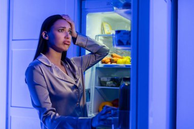 Pijamalı kafası karışmış genç bir kadın ürünlerle dolu açık buzdolabına bakıyor ve kafaya dokunuyor. Gecenin geç saatlerinde buzdolabının yanında duruyor.