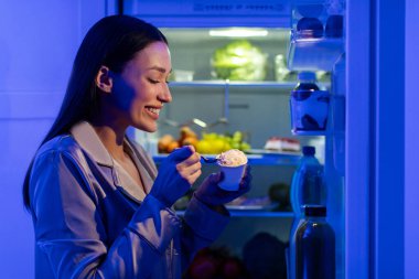 Buzdolabının yanında duran genç, aç bir kadın, tatlı dondurma yemekten zevk alıyor, gece geç saatlerde, yandan görüntülü çekim.