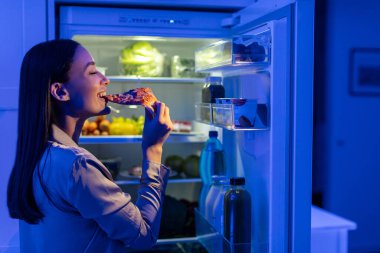 Pijamalı, pizza yiyen, geceleri açık buzdolabının yanında duran genç bir kadın. Diyeti bitir, kötü alışkanlık.