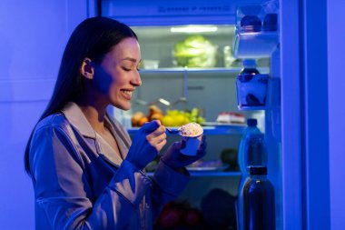 Lezzetli dondurma yiyen mutlu, aç bir kadın, geceleri açık buzdolabının yanında duruyor, diyetini bozuyor, yan görüş iğnesi vuruyor.