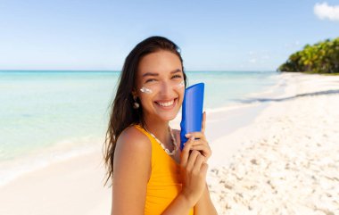 Güneş kremi tutan, nemlendirici güneş kremi süren, yüzüne krem süren, kumlu tropik plajda poz veren ve kameraya gülümseyen mutlu kadın.