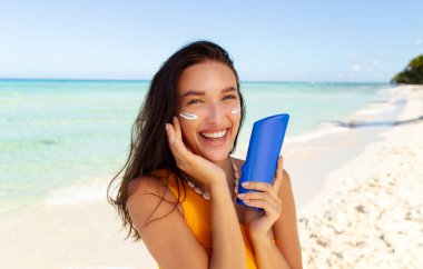 Yüzünde güneş kremi olan güzel bir kadın tropik plajda güneş kremi şişesi gösteriyor ve cildine nemlendirici losyon sürüyor. Cilt bakımı ve güneş koruma kavramı