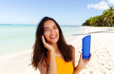 Mutlu kadın güneş kremi sürüyor şişe gösteriyor, yaz tatilinde kumlu sahilde duruyor, kameraya gülümsüyor.