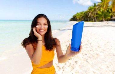 Güneş kremi şişesi gösteren, ultraviyole koruma için yüzüne losyon süren, kumlu tropik plajda duran, tasarım için modelleme yapan bir kadın.